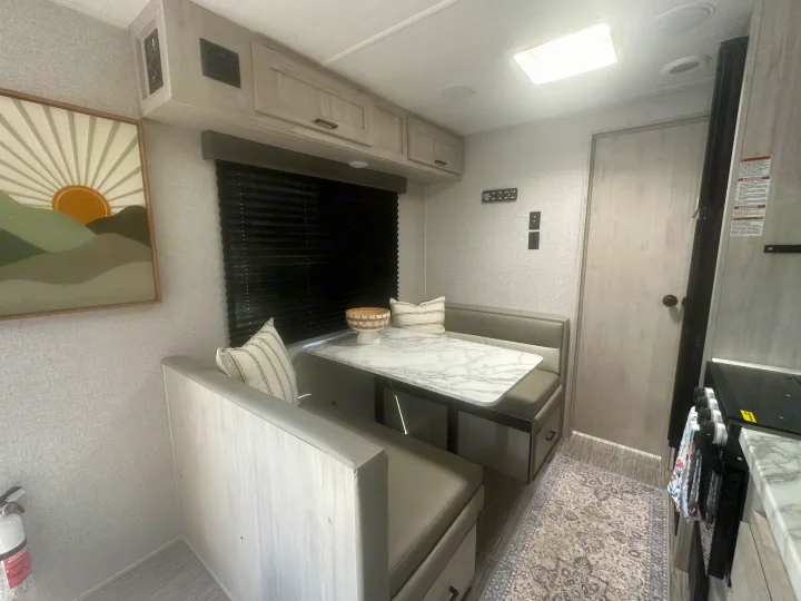Bright RV rental interior: dinette with modern table, comfy benches, window with blinds, cabinets, and kitchen.. East to West Silver Lake 2023