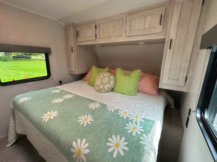 Cozy RV rental bedroom with a comfy bed, daisy-patterned blanket, overhead storage, and an outdoor view through the window.. East to West Silver Lake 2023
