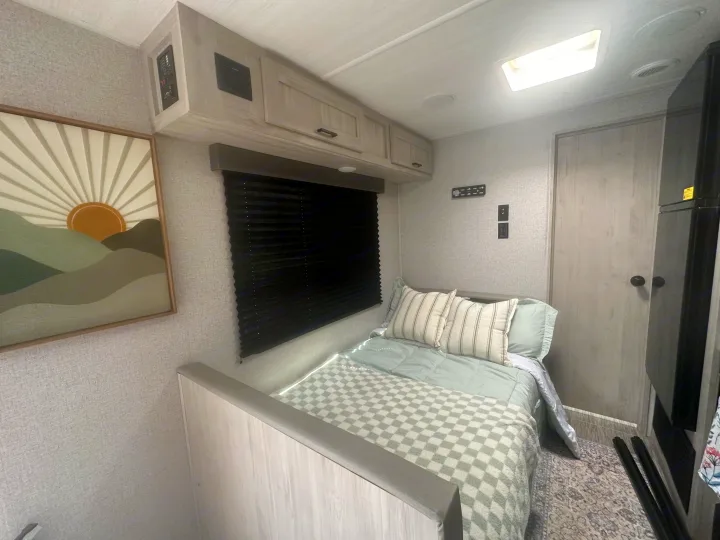 Modern RV rental interior: a clean bedroom with a cozy bed, overhead cabinets, and mountain art. Ideal for your next trip.. East to West Silver Lake 2023