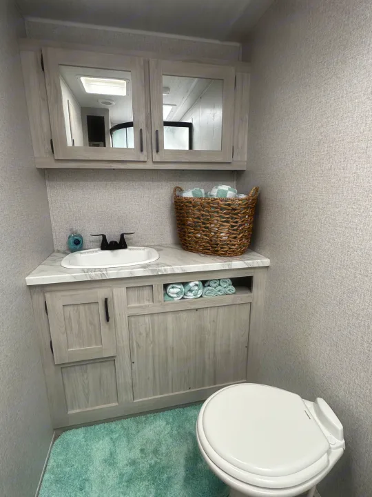 Inside an RV rental: a compact bathroom featuring a light wood vanity, white sink, toilet, and organized towel storage.. East to West Silver Lake 2023