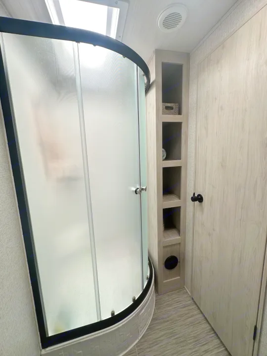 Inside an rv rental, featuring a modern bathroom with a curved frosted glass shower, built-in shelving, and light wood finishes.. East to West Silver Lake 2023