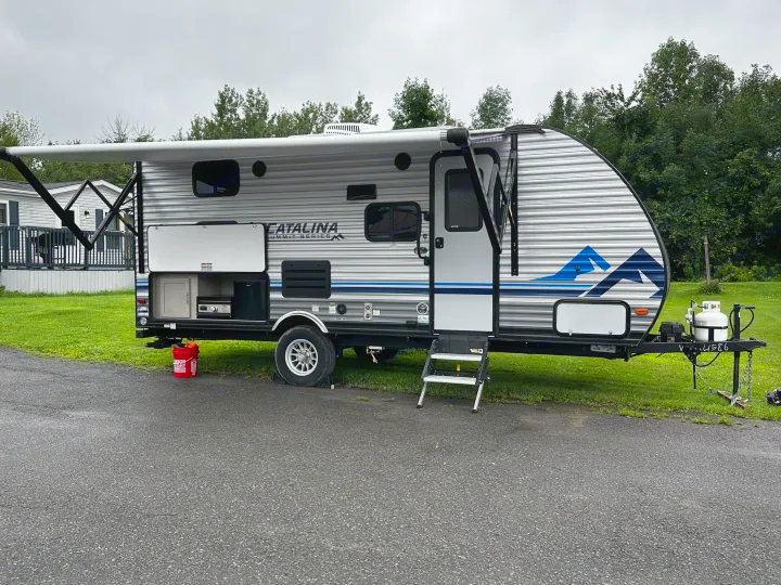 This RV rental features a modern travel trailer with an outdoor kitchen and extended awning, parked at a scenic campsite.. Coachmen Catalina 2022