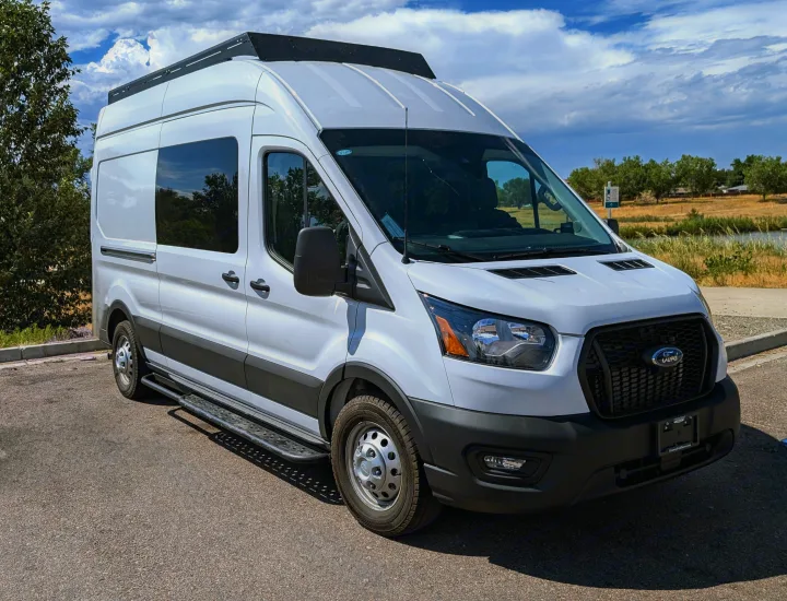 White Ford Transit rv rental van with side steps & roof rack, parked by trees & water under a cloudy sky.. Ford Van 2024