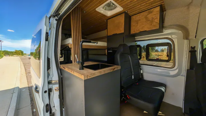 Experience this rv rental campervan interior: kitchen with sink, comfy seating, and elevated bed. Features wood ceilings, storage, and natural light.. Ford Van 2024