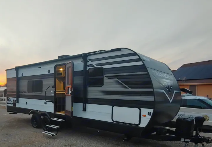 Modern gray and white Transcend RV rental parked on gravel with door open, illuminated inside, at sunset.. Grand Design Transcend Xplor 2022