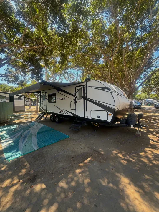 White RV rental parked at a shady campground site with an extended awning and picnic table.. Prime Time Tracer 2021