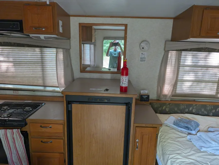 Inside an RV rental: a compact kitchen with stove & Dometic fridge, bed, cabinets, and safety devices. Mirror reflects outdoors.. Hi Lo 2307c 2007