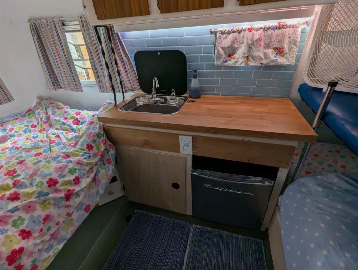 Cozy RV rental interior with a kitchenette, bed, and fan. Features a wooden counter, blue tile, and colorful bedding.. Boler Boler 1300 1974