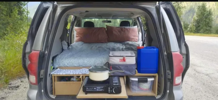 Inside a parked car, a cozy camping setup with a bed, portable stove, cooler, & storage for your next rv rental adventure.. Dodge Grand Caravan 2014