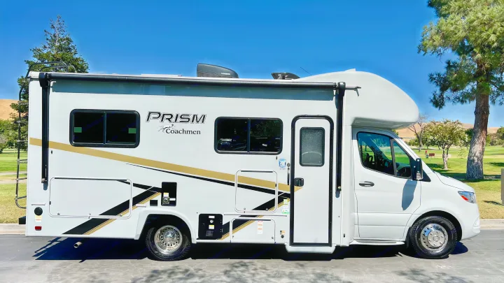 This Coachmen Prism RV rental, a white motorhome with gold & black stripes, is parked on asphalt with a sunny outdoor background.. Coachmen Prism 2024