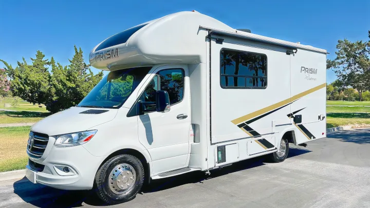 Modern white Class C RV rental, a Coachmen Prism motorhome, parked on asphalt with a clear sky and trees in the background.. Coachmen Prism 2024