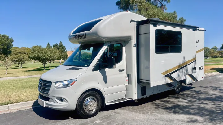 Bright white Prism RV rental, a sleek motorhome, parked on a sunny day near a golf course with trees and hills.. Coachmen Prism 2024