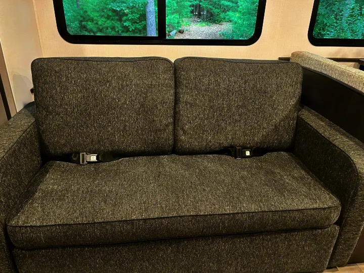 Grey couch with seatbelts in an RV rental, offering safe seating with views of trees.. Forest River Solera 27 DSE 2023