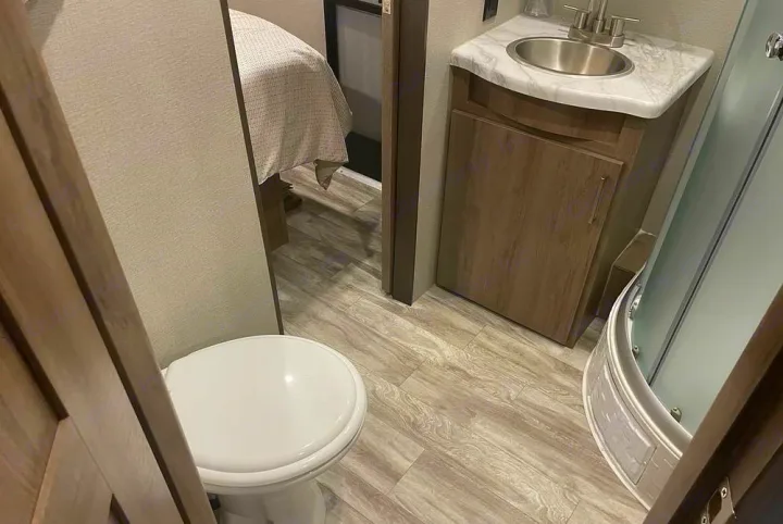 Inside an RV rental: Compact bathroom features a white toilet, vanity with sink, and curved shower, leading to the bedroom.. Grand Design Imagine 2023