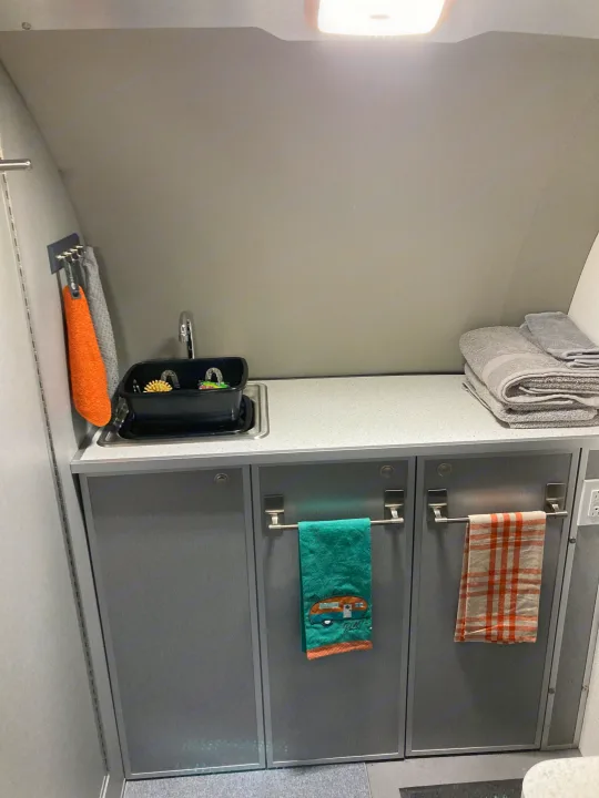 RV rental interior showing a small kitchen with a sink, dish rack, stacked towels, and cabinet storage.. Helio Helio Trailer 2023
