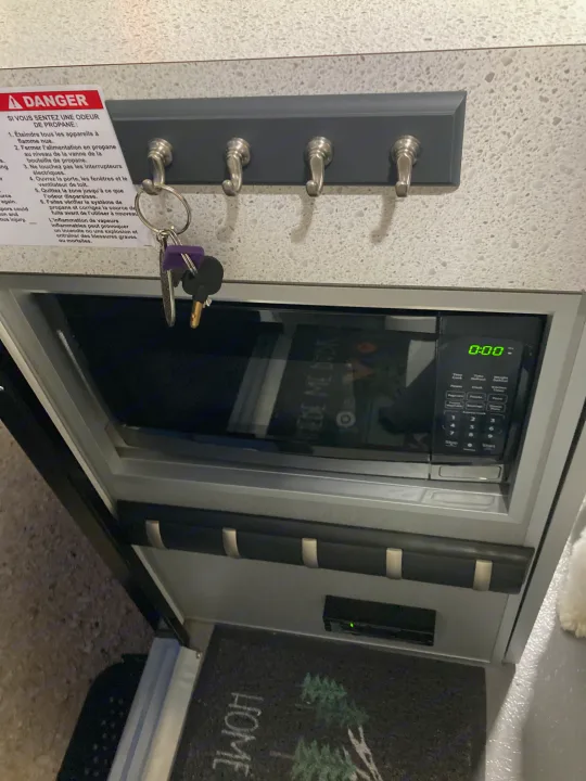 RV rental interior: A French propane safety sign, key hooks with purple keys, and a microwave set to "0:00.". Helio Helio Trailer 2023