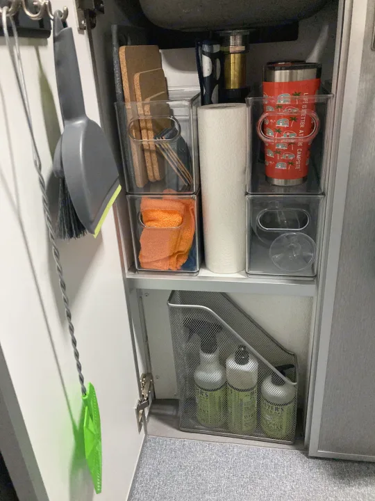 RV rental under-sink storage: organized cleaning supplies, towels, & mugs in clear bins & a mesh holder.. Helio Helio Trailer 2023