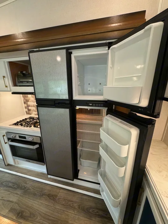 This RV rental kitchen features an open, empty fridge and freezer in a compact space, next to a stove, oven, and toaster oven.. Prime Time Crusader 2021