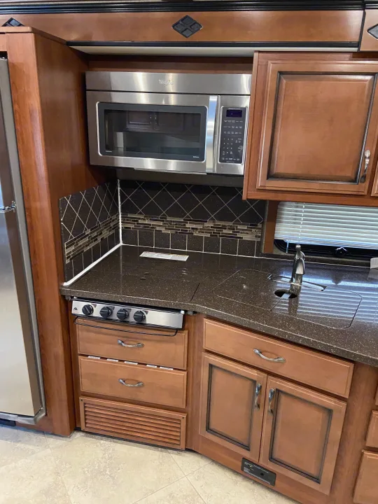 RV rental kitchen features a stainless microwave, stovetop, sink, and wood cabinets with dark speckled counters.. Fleetwood Other 2015