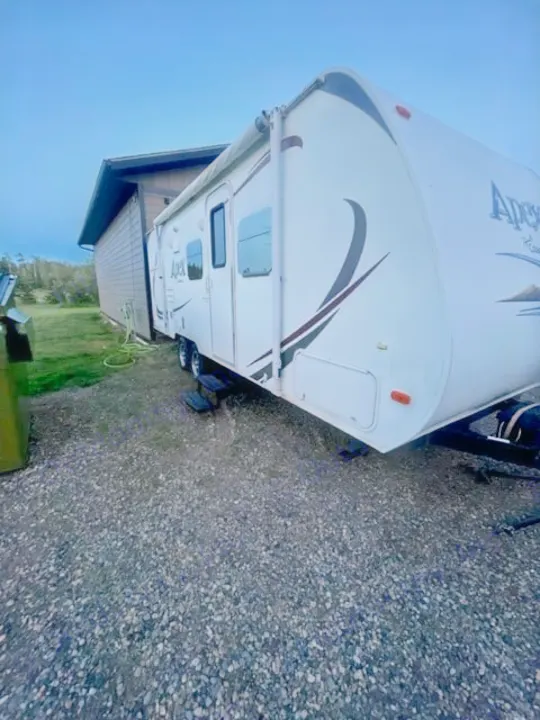 White Apex travel trailer RV rental parked on gravel beside a building, with its awning slightly extended.. Coachmen Apex 2013