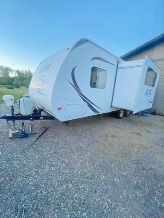 White Apex travel trailer with extended slide-out. Explore in this comfortable RV rental, parked on a gravel surface.. Coachmen Apex 2013
