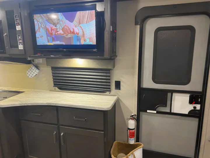 RV rental interior: A compact kitchen with a TV, dark cabinets, and a fire extinguisher mounted by the entry door.. Grand Design Transcend Xplor 2022