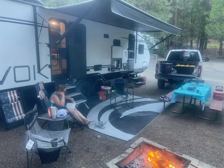 Enjoying an RV rental adventure: a travel trailer parked at a wooded campsite with a fire pit & outdoor living area.. Travel Lite Evoke 2020