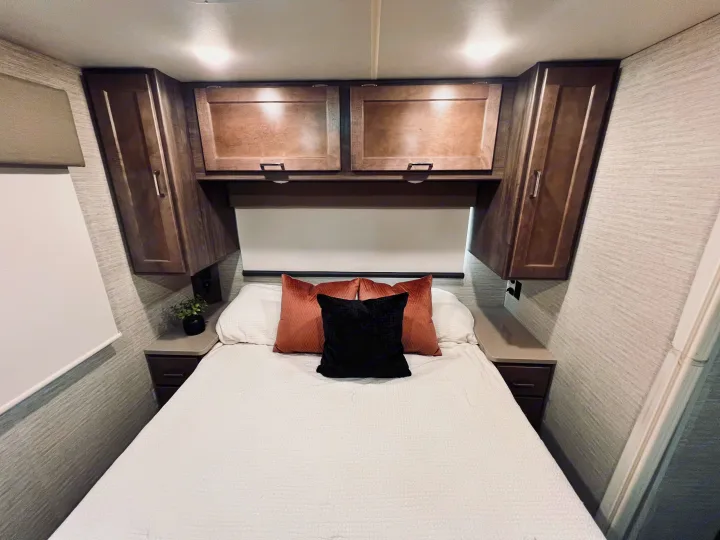 Cozy RV rental bedroom features a bed with pink & black pillows, overhead cabinets, and compact nightstands.. Winnebago Minnie Winnie 2022