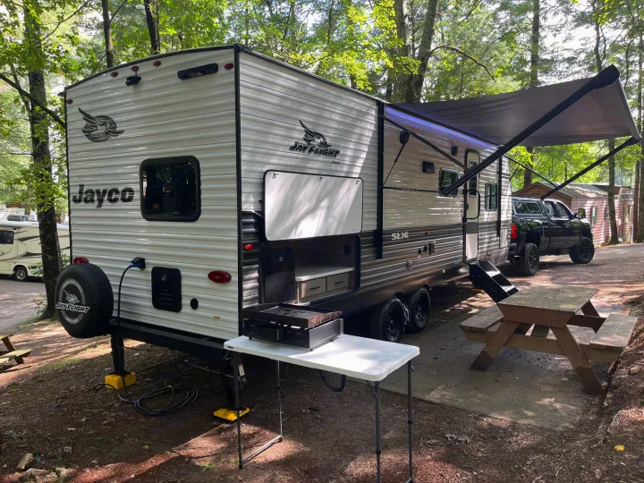 An RV rental in a wooded campsite features a Jayco travel trailer, outdoor kitchen, and extended awning.. Jayco JayFlight SLX 2024