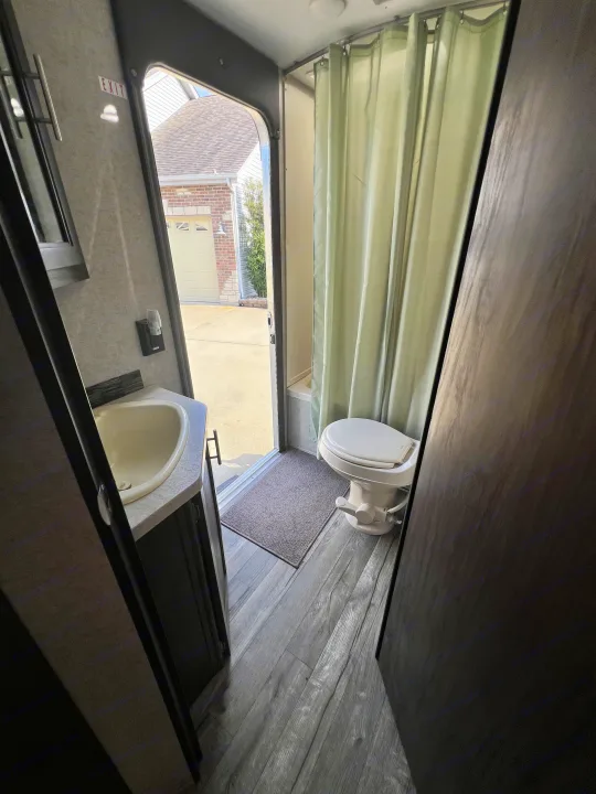 This RV rental bathroom features a compact sink, toilet, and shower with a light green curtain. The door is open to the outside.. Forest River Cherokee Grey Wolf 2019