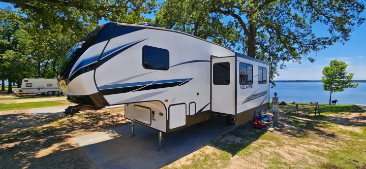 Spacious fifth-wheel RV rental parked lakeside at a scenic campground on a sunny day, ready for your travel adventures.. Crossroads Volante 2023