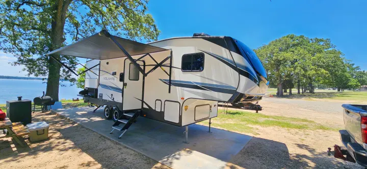 Lakeside RV rental campsite: A modern fifth-wheel camper with an extended awning by a lake, picnic table, and grill.. Crossroads Volante 2023
