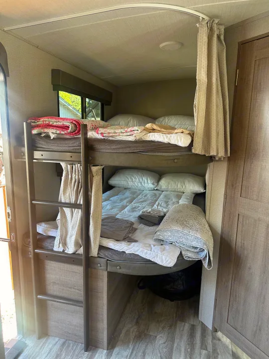 RV rental sleeping area: Two bunk beds, upper has pink quilt, lower has gray quilt. Ladder & privacy curtains are visible.. Grand Design Imagine 2020