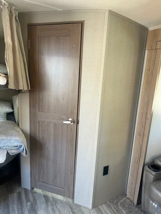 Interior of an RV rental: a wood-look door, light textured walls, and wood-patterned flooring. A bed and curtain are on the left.. Grand Design Imagine 2020