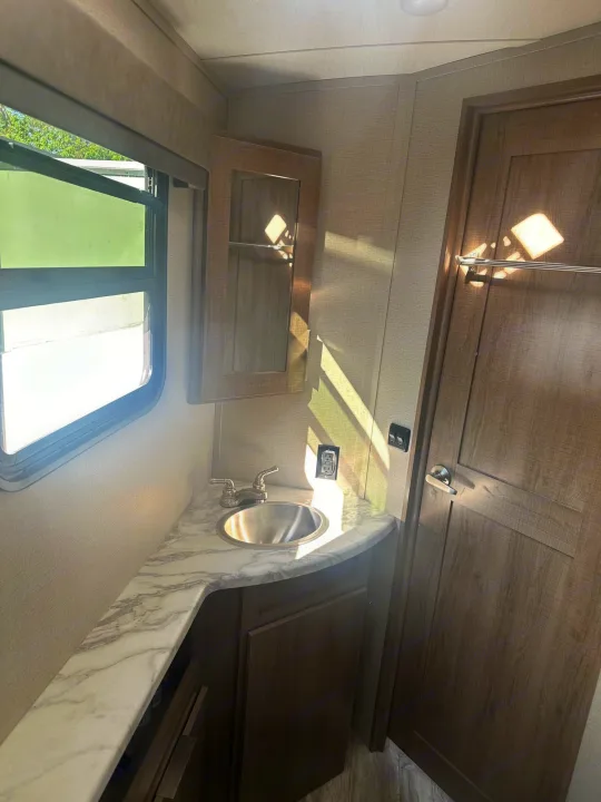 Compact RV rental bathroom features a corner sink, vanity, mirrored cabinet, bright window, and wooden door.. Grand Design Imagine 2020