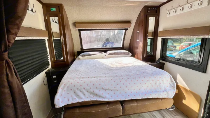 Interior of an RV rental bedroom showing a bed with a white and pink polka dot blanket, windows, and side cabinets.. Jayco White Hawk 2018