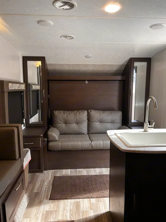 View of an RV rental interior featuring a gray sofa, dark wood cabinetry, kitchen sink, and wood-look flooring.. Forest River Wildwood Cruise Lite 2019