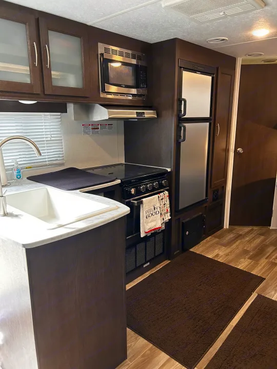 RV rental kitchen: Dark wood-look cabinets, stainless steel microwave, stove, and refrigerator in a compact cooking area.. Forest River Wildwood Cruise Lite 2019