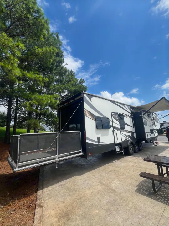 White & black XLR toy hauler RV rental parked with its rear patio deployed at a sunny campsite, amidst tall pine trees.. Forest River XLR Thunderbolt Toy Hauler 2015