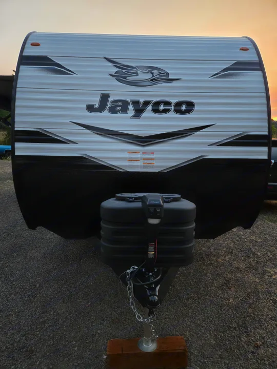 Front view of a white and black Jayco RV rental, featuring its logo, warning labels, and hitch on gravel.. Jayco Jay Flight SLX 2024