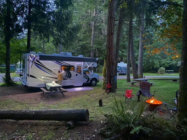 A white RV rental is set up at a forest campsite, complete with a picnic table and glowing campfire.. Thor Four Winds 2021