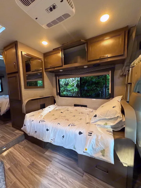 Cozy RV rental bedroom with patterned bedding, overhead cabinets, and a window view.. Thor Four Winds 2021
