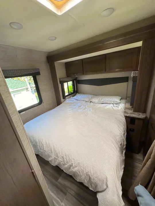 Spacious RV rental bedroom featuring a large bed with striped bedding, side windows, and overhead cabinets for comfortable mobile living.. Thor Four Winds 2021