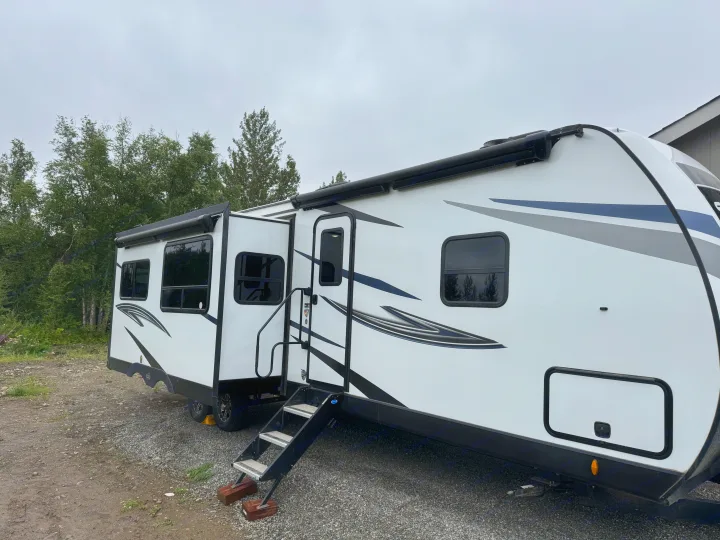 This white RV rental, a modern travel trailer with a slide-out, is parked on gravel amidst trees, ready for camping.. Venture RV 327VIK 2024