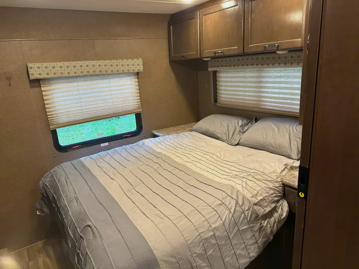 RV rental bedroom: a cozy bed with gray bedding, overhead cabinets, and a window with an "EXIT" sign in this compact space.. Thor Four Winds 2019