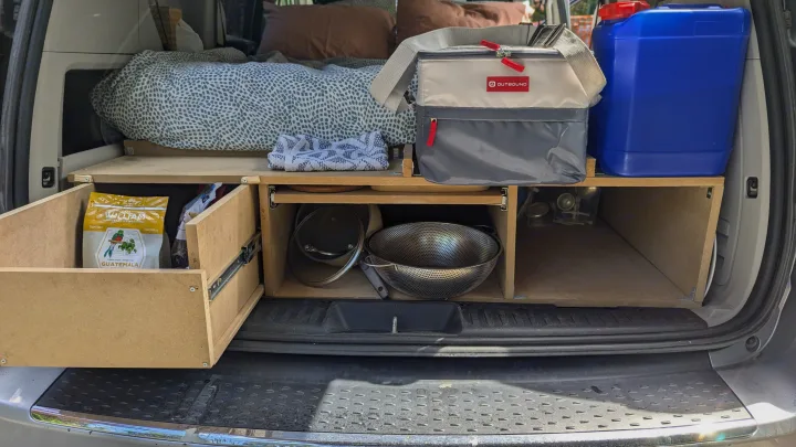 Compact rv rental kitchen & storage setup in a converted van. Features custom wooden shelves, pull-out drawer, cooler, & water jug.. Dodge Grand Caravan 2014