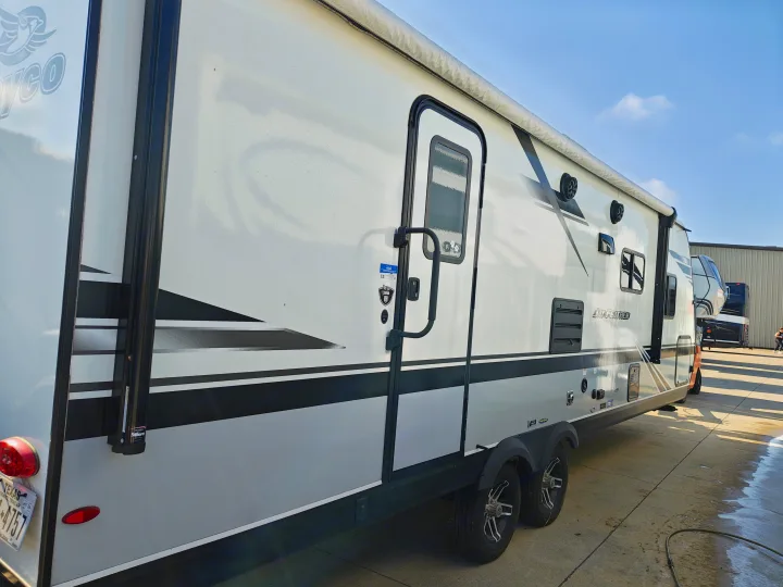 Side of a white and black travel trailer, featuring an awning and main door. Great for an RV rental, parked on concrete.. Jayco JAY FEATHER 26RB 2021