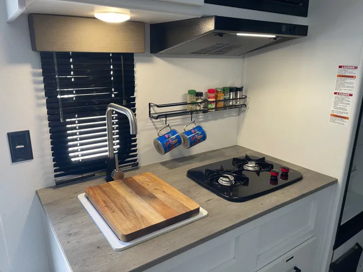 RV rental kitchen: gray countertop, black stove, sink with cutting board, spice rack, mugs, and window with blinds.. Keystone RV Hideout 2021