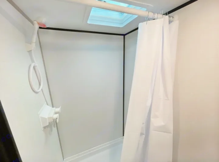Compact RV rental shower: a white stall with handheld showerhead, skylight, and drawn white curtain.. Coleman 17B 2023