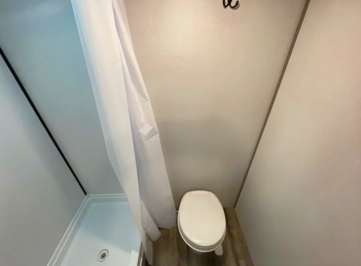 Compact RV rental bathroom featuring a white shower with curtain on the left and a toilet on wood-look flooring.. Coleman 17B 2023